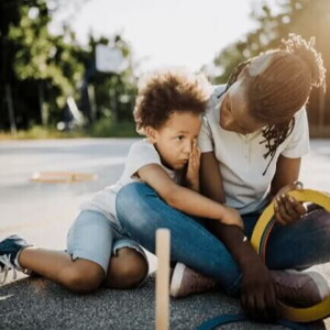 Mãe e filho, uma criança de 6 anos, estão sentados no chão de um espaço ao ar livre. A mãe olha para o filho, que o abraça.