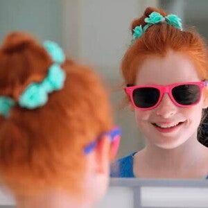 Menina sorri para a própria imagem no espelho. Se trata de uma criança de cerca de 9 anos usando óculos escuros com armação rosa.