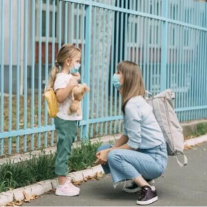Uma mãe está agachada em frente a sua filha, perto de um portão. A menina está de mochila, olhando para a mãe e segurando um urso de pelúcia.