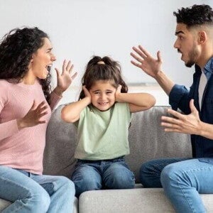 Pais discutindo sentados no sofá enquanto a criança, entre eles, cobre os ouvidos, demonstrando desconforto diante do conflito familiar.