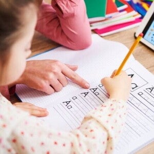 Uma mão adulta aponta para um caderno quadriculado onde uma criança está praticando a escrita das letras "A" e "a" com um lápis.