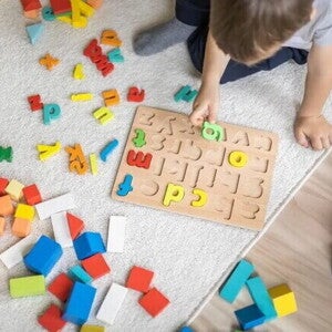 Em um piso de madeira com um tapete cinza, está sentado um menino,vestindo uma camiseta branca. Ele está brincando com um tabuleiro de encaixar letras, enquanto segurando uma delas. Há letras e outras peças coloridas espalhadas ao seu redor. Em um piso de madeira com um tapete cinza, está sentado um menino,vestindo uma camiseta branca. Ele está brincando com um tabuleiro de encaixar letras, enquanto segurando uma delas. Há letras e outras peças coloridas espalhadas ao seu redor.