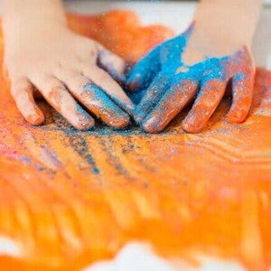 Imagem das mãos de uma criança, sentindo a textura de um glitter e de tintas. Ao fundo, um cartaz branco com algumas pinturas na cor laranja, rosa e glitter azul. Imagem das mãos de uma criança, sentindo a textura de um glitter e de tintas. Ao fundo, um cartaz branco com algumas pinturas na cor laranja, rosa e glitter azul.