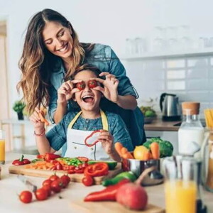 Uma mãe e sua criança estão na cozinha, com diversos alimentos sobre uma bancada. Ela segura dois tomates pequenos na altura dos olhos da criança e ambas estão sorrindo