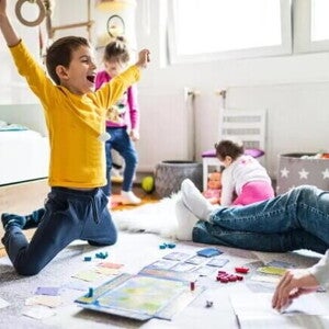 Sala com um adulto, onde só é possível ver suas pernas usando calça jeans e meia. Na mesma sala, estão três crianças. Uma garota com cerca de 1 ano de idade em segundo plano, branca, de cabelos curtos, de costas brincando com uma boneca. Outra garota com cerca de quatro anos, branca, cabelos loiros presos, calça jeans e blusa de manga comprida. Ela parece estar feliz dançando. A criança em destaque é um menino, branco, com cabelos curtos, de braços abertos comemorando uma vitória em um jogo 