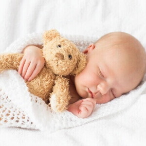 Bebê dormindo tranquilamente enrolado em uma manta branca e abraçado em um urso pequeno de pelúcia.