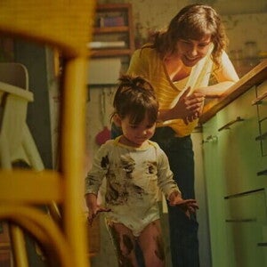 Mãe vestida com uma blusa amarela, encostada na bancada da cozinha, observa, sorridente, a filha que está suja com tinta nas mãos, nas pernas e na roupa Mãe vestida com uma blusa amarela, encostada na bancada da cozinha, observa, sorridente, a filha que está suja com tinta nas mãos, nas pernas e na roupa