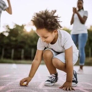 Imagem de uma criança ao centro brincando de uma brincadeira africana no chão. Ao fundo seus pais torcem por ela