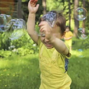 Um menino sorri com as mãos para o alto enquanto passa por bolhas de sabão em um ambiente externo arborizado, com outro menino ao fundo. A foto faz alusão ao tema de brincadeiras ao ar livre. 