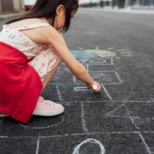 Uma menina de vestido e tênis está agachada na rua desenhando uma amarelinha no chão. A foto faz alusão ao tema de brincadeiras de rua Uma menina de vestido e tênis está agachada na rua desenhando uma amarelinha no chão. A foto faz alusão ao tema de brincadeiras de rua