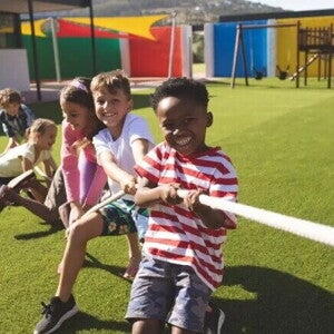 Em um parque ao ar livre, três meninos e duas meninas estão se divertindo em uma brincadeira de cabo de guerra. As crianças, sorridentes, estão puxando a corda.