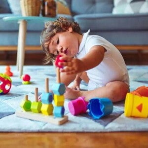 Um bebê está sentado no tapete de uma sala de estar mexendo com brinquedos de empilhar e montar, que é uma das brincadeiras que estimulam o desenvolvimento infantil Um bebê está sentado no tapete de uma sala de estar mexendo com brinquedos de empilhar e montar, que é uma das brincadeiras que estimulam o desenvolvimento infantil