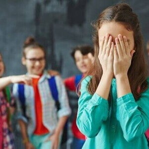 Grupo de crianças apontando e rindo de uma menina que está cobrindo o rosto com as mãos