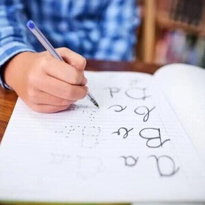 Imagem de um menino segurando uma caneta azul. Em um caderno ele vai ligando os pontinhos para formar as palavras: a,b,c e d. A foto faz alusão ao tema caligrafia. Imagem de um menino segurando uma caneta azul. Em um caderno ele vai ligando os pontinhos para formar as palavras: a,b,c e d. A foto faz alusão ao tema caligrafia.