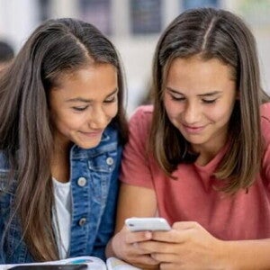 Duas meninas sentadas uma ao lado da outra na sala de aula, sorrindo e olhando para um celular