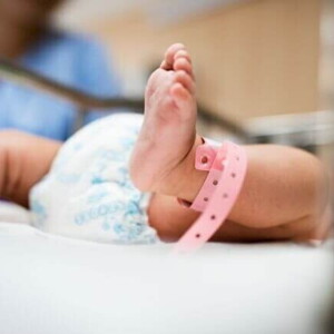 Close de um bebê recém-nascido deitado em um berço hospitalar, com uma pulseira rosa de identificação no tornozelo, simbolizando cuidados neonatais e saúde infantil.