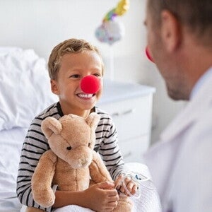 Um menino sorridente com um nariz de palhaço vermelho e segurando um ursinho de pelúcia está sentado em uma cama de hospital, olhando para um médico de costas para o observador. O médico também usa um nariz de palhaço vermelho.