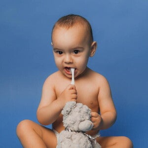 Bebê sentado com uma escova de dentes na boca, segurando um urso de pelúcia