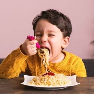 Criança sentada à mesa, comendo um prato de macarrão, em ambiente interno com parede rosa e planta ao fundo. Criança sentada à mesa, comendo um prato de macarrão, em ambiente interno com parede rosa e planta ao fundo.
