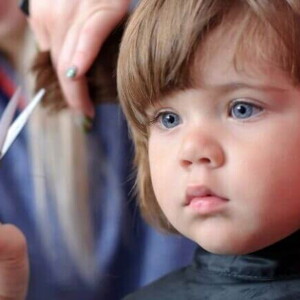 Uma menina recebendo um corte de cabelo. Ao seu lado, a mão de uma mulher segura uma mecha do seu cabelo, enquanto outra mão segura uma tesoura pequena
