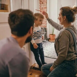A mãe está ajoelhada checando o crescimento no filho, que está encostado na parede. Ela segura um lápis sobre a cabeça da criança para marcar a altura. O pai olha para a cena, um pouco mais distante. 