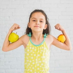 Menina sorridente fazendo pose de força com os braços flexionados, segurando uma maçã em cada braço, em frente a uma parede branca. Menina sorridente fazendo pose de força com os braços flexionados, segurando uma maçã em cada braço, em frente a uma parede branca.