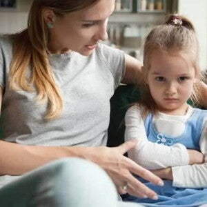 Mãe e filha sentadas no sofá da sala, mãe abraça a criança e conversa, criança parece estressada com os braços cruzados e olhar distante Mãe e filha sentadas no sofá da sala, mãe abraça a criança e conversa, criança parece estressada com os braços cruzados e olhar distante
