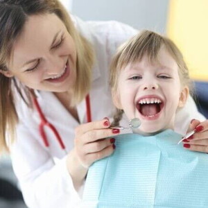 Criança sentada com a boca aberta, ao lado de uma dentista que está em pé, sorrindo e segurando aparelhos odontológicos.