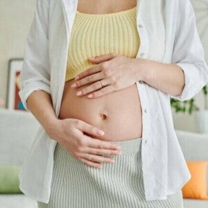 Uma mulher grávida em pé, com as duas mãos sobre a barriga.