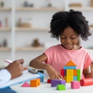 Uma menina sorrindo e organizando blocos coloridos, ao lado de uma mulher sentada com uma ficha na mão.
