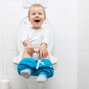 Uma criança sentada em um vaso sanitário com os shorts abaixados, rindo e olhando para cima, em um banheiro.