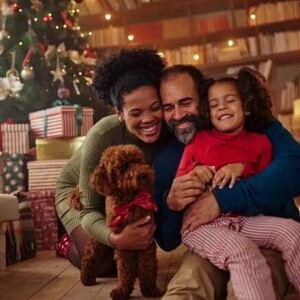 Homem e mulher abraçados com a filha e um cachorrinho no chão, na sala de casa em frente a uma árvore de natal