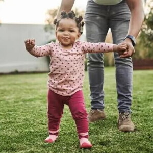 Imagem de um bebê cuja criação segue a abordagem de Emmi Pikler e que está brincando em um quintal supervisionado pela mãe que está lhe dando a mão.