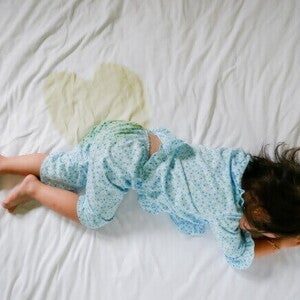 Criança deitada de bruços em uma cama com lençol branco, com uma mancha de xixi visível ao lado, vestindo pijama azul claro com estampa. Criança deitada de bruços em uma cama com lençol branco, com uma mancha de xixi visível ao lado, vestindo pijama azul claro com estampa.