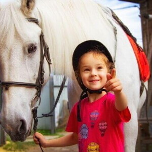 Menina sorrindo e apontando com o dedo, enquanto segura a rédea de um cavalo