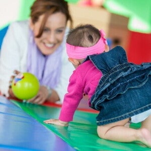 Um bebê engatinhando em direção a uma mulher sorridente, que observa o bebê com uma bola verde na mão. Um bebê engatinhando em direção a uma mulher sorridente, que observa o bebê com uma bola verde na mão.