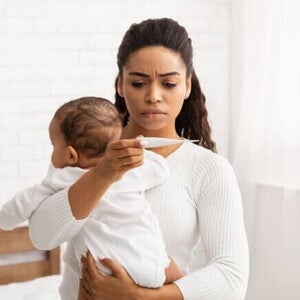 Uma mulher segura um bebê nos braços, com feição preocupada, enquanto verifica a temperatura do bebê com um termômetro.
