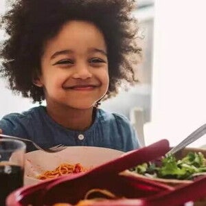 ferro na alimentacao infantil criança sentada a mesa no horário de almoço, sorrindo enquanto segura o talher na mão. Em sua frente estão os pratos com alimentos, incluindo uma salada verde, e há a presença de um adulto do outro lado da mesa.