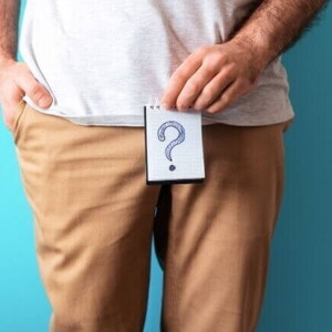 Homem de camiseta branca e calça marrom segurando um papel desenhado um ponto de interrogação em frente ao seu órgão genital