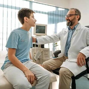 Imagem de um ambiente hospitalar em que um médico tranquiliza um paciente menino, ao colocar a mão no ombro dele. O médico está de jaleco branco, óculos e tem barba e o menino está vestindo uma camiseta azul Imagem de um ambiente hospitalar em que um médico tranquiliza um paciente menino, ao colocar a mão no ombro dele. O médico está de jaleco branco, óculos e tem barba e o menino está vestindo uma camiseta azul