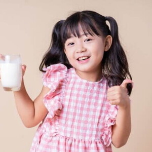 Menina sorrindo, vestindo um vestido xadrez rosa e branco, segurando um copo de leite e fazendo um joinha
