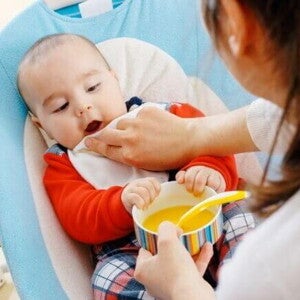 Bebê sentado em uma cadeirinha, com as mãos em um potinho de comida, sendo limpo por um adulto utilizando uma fralda de boca. Bebê sentado em uma cadeirinha, com as mãos em um potinho de comida, sendo limpo por um adulto utilizando uma fralda de boca.
