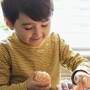 Um menino, vestido com camisa listrada, segura uma tangerina. Entrando no quadro há uma mão de adulto dando frutas para a criança.