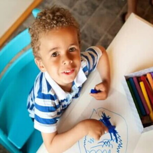 criança sentada em cadeira e mesa infantil com giz de cera azul em mão, desenhando em folha de papel, outras cores de giz disponíveis sobre a mesa