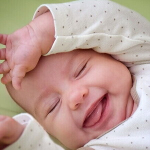 Bebê sorrindo de olhos fechados, com a mão perto do rosto, vestindo roupa branca com bolinhas cinzas. Bebê sorrindo de olhos fechados, com a mão perto do rosto, vestindo roupa branca com bolinhas cinzas.
