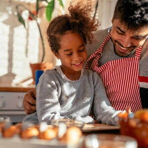 A relação de pai e filho é ilustrada por uma imagem onde a menina é abraçada pelo pai, que veste avental, enquanto eles olham para uma mesa de café da manhã com alguns alimentos. A relação de pai e filho é ilustrada por uma imagem onde a menina é abraçada pelo pai, que veste avental, enquanto eles olham para uma mesa de café da manhã com alguns alimentos.