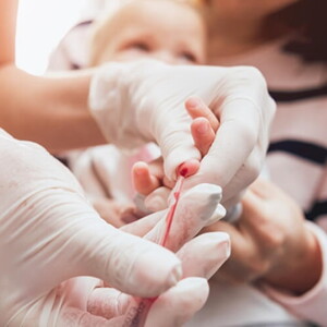 Mãe segurando bebê enquanto enfermeira colhe sangue para exame.