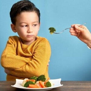 Uma mão estendendo um garfo com brócolis para um menino, que olha de forma irritada para o alimento.