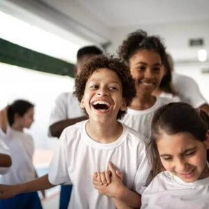 Imagem de um ambiente escolar com várias crianças vestidas de uniforme. O destaque está para um menino gargalhando, bem no centro da foto, de mãos dadas com uma menina Imagem de um ambiente escolar com várias crianças vestidas de uniforme. O destaque está para um menino gargalhando, bem no centro da foto, de mãos dadas com uma menina