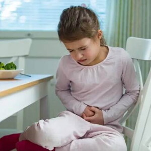 Menina com intoxicação alimentar, sentada à mesa de jantar, reagindo à dor, colocando as mãos na barriga Menina com intoxicação alimentar, sentada à mesa de jantar, reagindo à dor, colocando as mãos na barriga